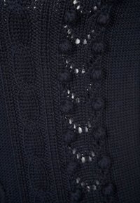 Marineblaues Stricktextil mit Kabelmustern, Quastendetails und Durchbrüchen, die ein strukturiertes und schichtartiges Erscheinungsbild schaffen.