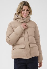 Veste rembourrée beige avec un col montant, une fermeture à pression, des poignets côtelés et deux poches avant. Texture lisse avec un design matelassé.
