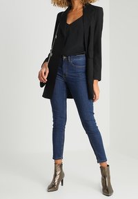 Kvinna klädd i svart kavaj, svart topp, blå skinny jeans, metalliska ankelstövlar och svart väska med kedja som står mot en enfärgad bakgrund.