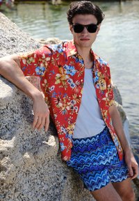 Jonge man met zonnebril, rood bloemenoverhemd en blauwe korte broek met patroon, leunend tegen een grote rots bij kalm water.