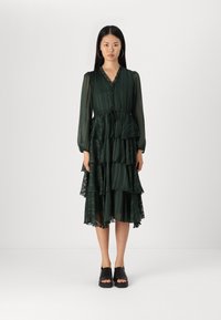 Grön lagerklänning med långa, genomskinliga ärmar, knappstängning fram, och ett bälte i midjan, med en volangkant och veckad textur.