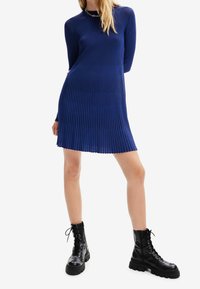 Vestito a maniche lunghe in maglia a coste di un blu profondo, con silhouette dritta e scollatura rotonda. Abbinato a stivali militari neri con lacci.