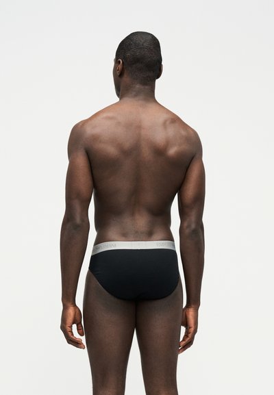 Άνδρας μοντέλο που στέκεται με την πλάτη, φορώντας μαύρα εσώρουχα με γκρι ζώνη μέσης, σε απλό γκρι φόντο.