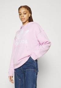 Rosa hettegenser laget av mykt stoff, med en stor hvit Adidas-logo og kengurulomme. Kombinert med mørke denimbukser.