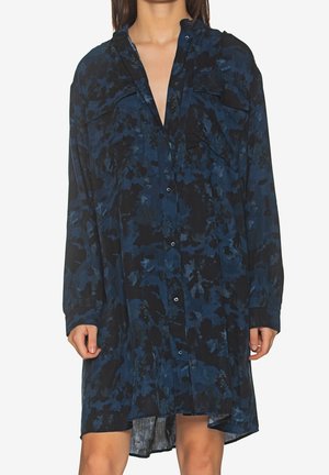 Femme portant une robe chemise ample à manches longues avec un motif abstrait bleu foncé et noir, debout les bras détendus.