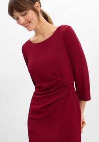Abito borgogna con scollatura rotonda e maniche a tre quarti. Presenta un tessuto arricciato in vita per un effetto plissè. Texture liscia.