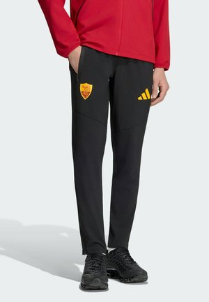 Persona con pantalones deportivos negros con logos amarillos de AS Roma y Adidas, chaqueta roja y zapatillas negras de correr, de pie con las manos parcialmente en los bolsillos.