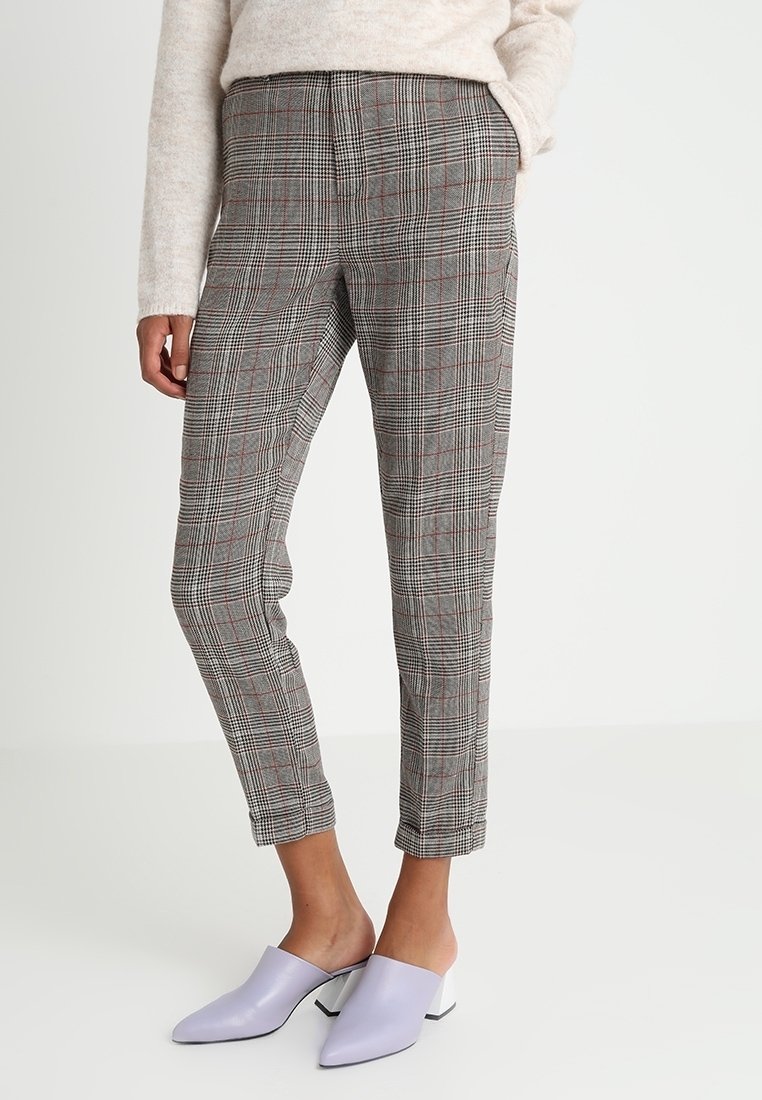 Pantalon à carreaux gris avec des accents rouges, coupe ajustée, taille haute et textures subtiles, associé à des mules violet clair.