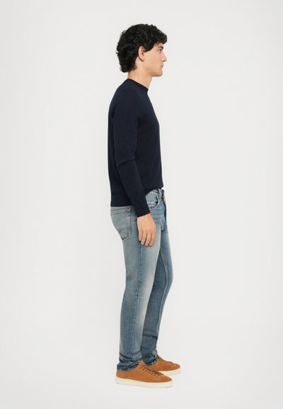 Man standing in profile wearing a navy sweater, light blue jeans, and brown suede sneakers against a plain white background.