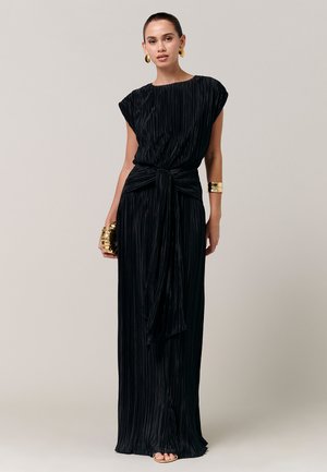 Frau steht in einem schwarzen, plissierten, bodenlangen Kleid mit gebundenem Taillenband, trägt ein goldenes Armreif, goldene Ohrringe und hält eine goldene Clutch.