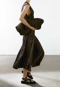 Brown pleated maxi dress with a high neckline and side cutout, held with one hand. Pair of black strappy sandals on feet.