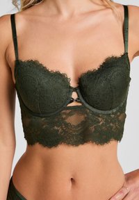 Donkergroene kanten bralette met onderdraadbekers, gekartelde randen en verstelbare dunne bandjes, gedragen op een blote torso.