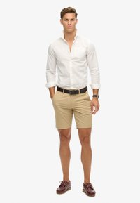 Hvid langærmet skjorte med oprullede ærmer, beige shorts, sort bælte og brune bådsko. Model står foran en neutral baggrund.