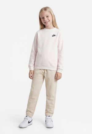 Jeune fille souriante, portant un sweat-shirt Nike rose pâle, un pantalon de survêtement beige, et des baskets blanches Nike, debout sur un fond blanc.