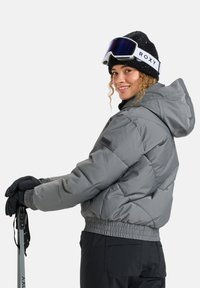 Giacca a vento grigia con cappuccio e motivo trapuntato, abbinata a guanti neri e un berretto nero. Presenta un design corto e una vita elasticizzata.