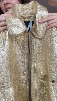 Osoba drži sjajnu zlatnu jaknu teksture, pokazujući njen ovratnik i unutarnju oznaku s velikim bijelim slovom na crnoj pozadini.