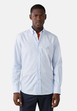 Hombre con una camisa abotonada de rayas azul claro sobre una camiseta naranja y pantalones oscuros, de pie frente a un fondo claro y sencillo.