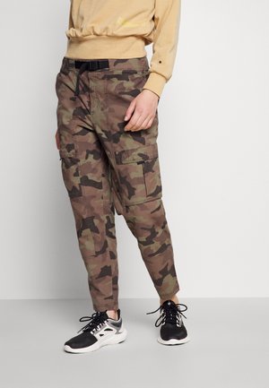 Persoon draagt bruine en groene camouflage cargobroek met zwarte riem en zwart-witte sneakers, staand tegen een effen muur.