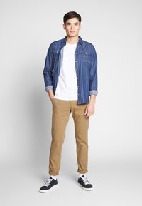 Jeanshemd met verticale blauwe strepen, witte t-shirt, beige chinos en zwarte sneakers met witte zolen. Casual pasvorm en opgerolde mouwen.