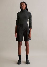 Charcoal-grijs geribde turtleneck, hoog-waist zwarte shorts met plooien, zwarte chunky loafers en zwarte enkelsokken tegen een lichte achtergrond.