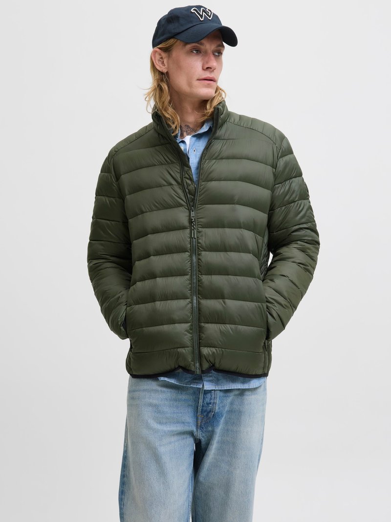 Geacă puffer de culoare verde măslinie, cu fermoar în față, guler înalt și design matlasat orizontal. Poartă o cămașă de culoare albastră deschisă și blugi.
