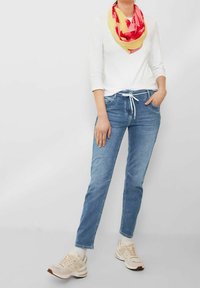 Jeans en denim bleu avec une taille ajustable à cordon, associés à un haut à manches longues blanc et une écharpe multicolore. Des baskets beige complètent la tenue.