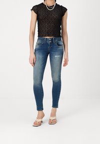 Top corto de encaje negro con mangas cortas con volantes, combinado con jeans ajustados azules con un acabado desgastado y detalles desgastados. Sandalias de color claro.