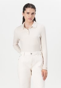 Ribbet langermet polo-skjorte i lys beige, med kragehals og knappestolpe. Kombinert med kremfargede høyt midje bukser.