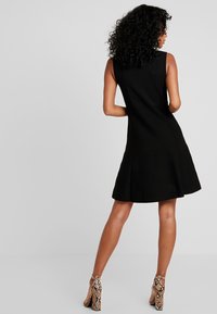 Femme aux cheveux bouclés portant une robe noire sans manches et des chaussures à talons hauts en imprimé serpent, se tenant devant un fond uni.