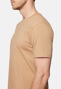 Beżowa koszulka z krótkim rękawem, wykonana z miękkiej bawełny, z okrągłym dekoltem oraz dopasowanym krojem, z widocznym przeszyciem na rękawach.