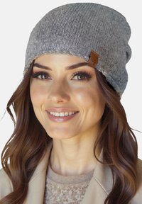 Kvinde med bølget brunt hår iført en grå strikket hue og beige jakke, smilende blidt mod en ensfarvet baggrund.