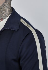 Mornarska zip-up jakna s preklopljenim ovratnikom, kremastim bočnim trakama i brendiranim oznakom na ramenu. Glatka tekstura tkanine.
