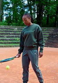 Zöld pulóver "BJÖRN BORG" felirattal, szürke sportnadrág és egy teniszütő. A felület narancsszínű agyag, a háttérben fákkal.