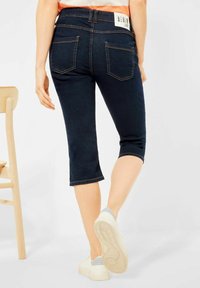 Tummansiniset denimin caprihousut, joissa on takataskut, tikatut yksityiskohdat ja käännetty lahje; yhdistetty vaaleisiin tennareihin.