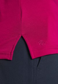 Magenta puuvillane T-särk, millel on ventileeritud äär ja küljes tikand "RL" logo, koos tumedate riidest pükstega.