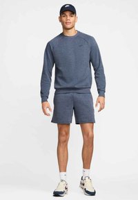 Sudadera deportiva azul marino con cuello redondo, combinada con pantalones cortos a juego. Acompañada de una gorra azul marino y zapatillas crema con detalles oscuros.