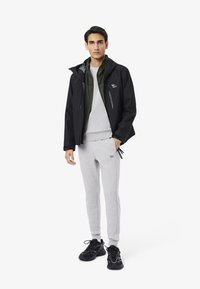 Cazadora negra con cremalleras, capa interior verde, sudadera gris y pantalones de chándal grises. Zapatillas negras completan el conjunto.