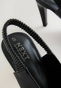 Mustat korkeakorkoiset sandaalit, joissa on teksturoitu stiletto, punottu hihna ja sileä nahkapinta. Logo painettuna sisäpohjaan.