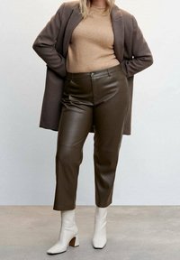 Braune Lederhosen mit schmaler Passform, mittelhohem Bund und dezentem Glanz, kombiniert mit einem hellbraunen Pullover und einem langen braunen Mantel. Weiße Stiefel mit Absatz vervollständigen den Look.