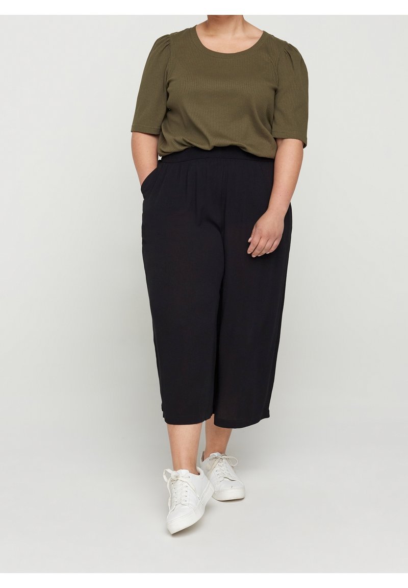 Personne portant un haut à manches courtes vert olive, un pantalon noir large et court, et des baskets blanches, tenant les mains partiellement dans les poches.