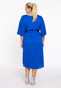 Blauwe, middelange jurk met driekwart mouwen, een strikceintuur en een gladde textuur. Gearchiveerd met zilveren enkelsandalen met een bandje.