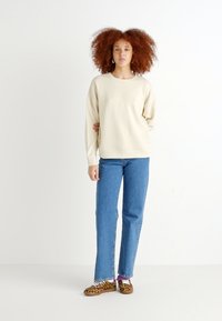Helles beiges Fleece-Sweatshirt mit Rundhals, kombiniert mit blauen gerade geschnittenen Jeans. Dazu passen Schuhe mit Leopardenmuster zum Hineinschlüpfen.