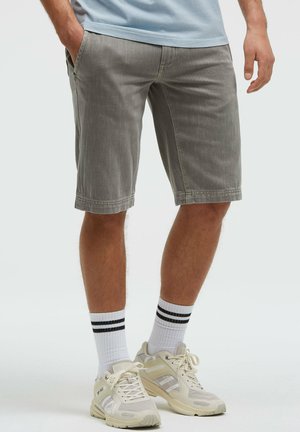 Mann trägt graue knielange Shorts, weiße Sportsocken mit schwarzen Streifen und beige Sportschuhe, steht mit entspannten Händen.