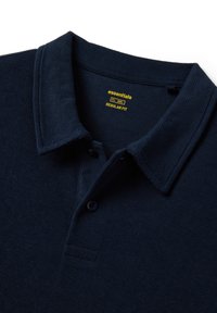 Polo de punto azul marino con cuello y dos botones, etiquetado como "essentials" en texto amarillo para un ajuste regular.
