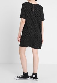 Schwarzes Polka-Dot-Kleid mit kurzen Ärmeln, einem runden Ausschnitt und einem Reißverschlussdetails im Rücken. Kombiniert mit schwarzen High-Top-Sneakern.