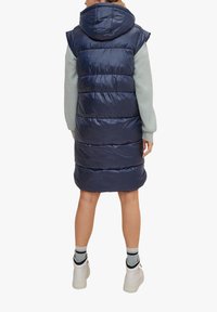 Marineblaue gesteppten Daunenweste mit Kapuze, mit horizontalem Steppmuster und glänzender Textur. Wird über einem hellgrünen Pullover getragen und mit weißen Schuhen kombiniert.