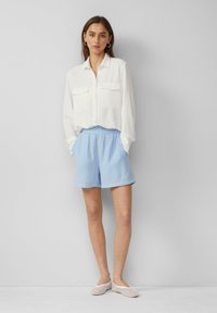 Chemise blanche à boutons avec deux poches poitrine, assortie à un short en tissu texturé bleu clair et des chaussures slip-on en mesh. Design simple et décontracté.
