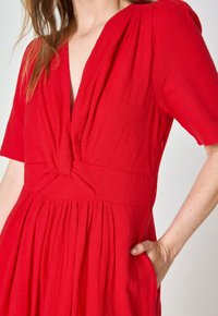 Red dress in lightweight fabric with a V-neck, pleated skirt, and knotted waist detail, featuring short sleeves and side pockets.