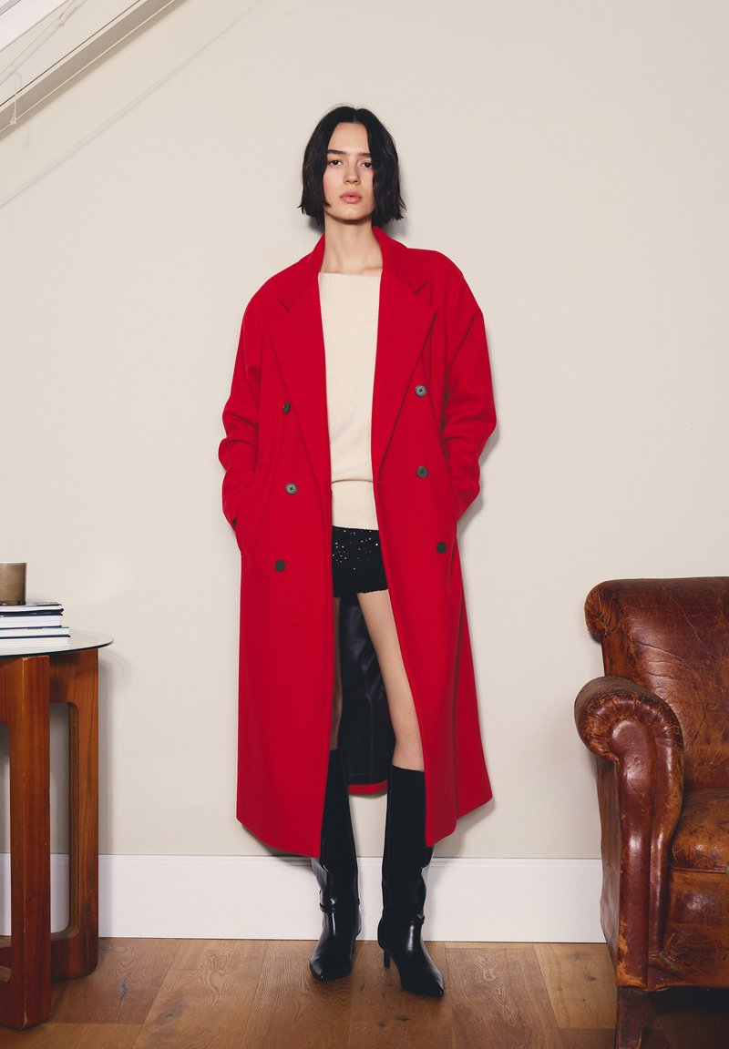 Červený dvojradový vlnený kabát s veľkými klopami, bočnými vreckami, nosený cez béžový sveter a čierne šortky, doplnený čiernymi čižmami po kolená.