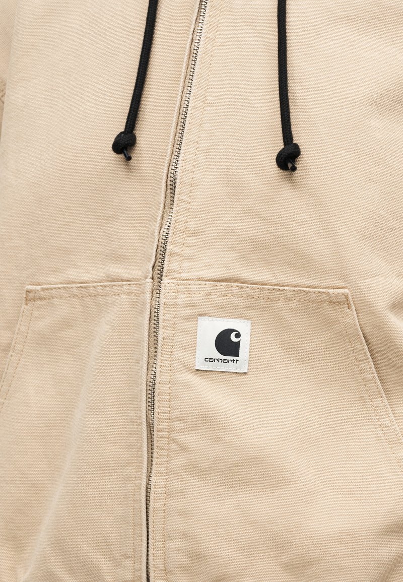 Sweat à capuche beige zippé avec cordons noirs et une étiquette carrée blanche présentant un logo noir et le nom de la marque sur la poche avant.